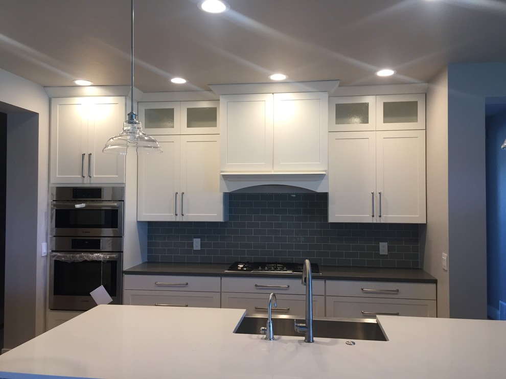 Example of a transitional l-shaped medium tone wood floor open concept kitchen design with a farmhouse sink, shaker cabinets, white cabinets, quartz countertops, gray backsplash, subway tile backsplash, stainless steel appliances and an island