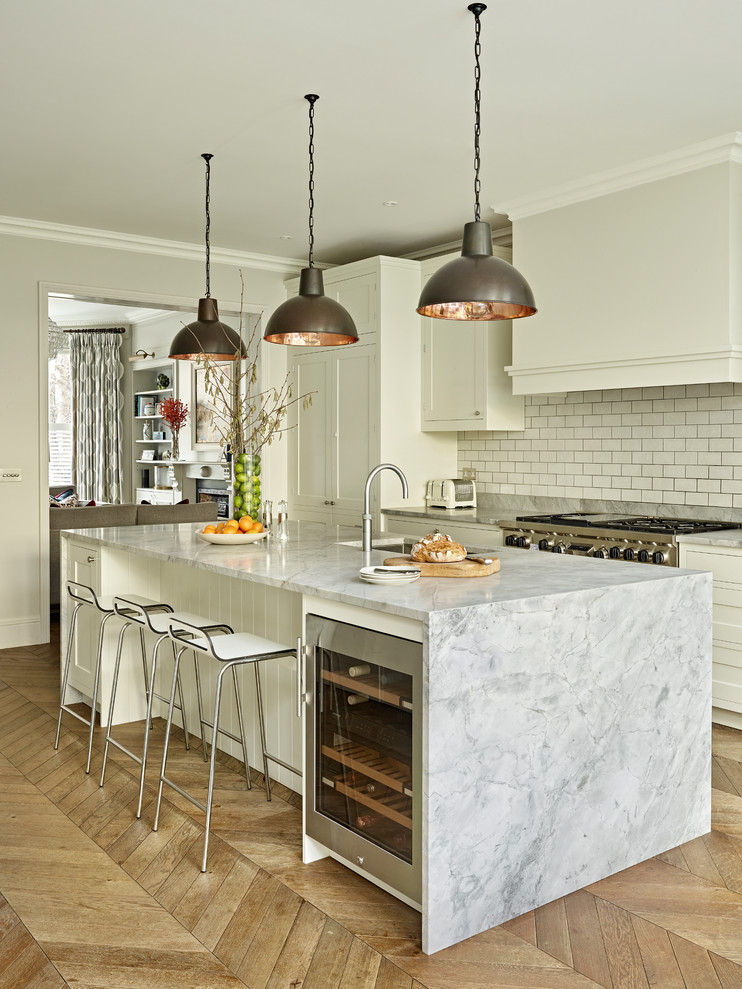 Inspiration for a transitional galley kitchen in London with an undermount sink, shaker cabinets, white cabinets, white splashback, subway tile splashback, stainless steel appliances, medium hardwood floors, with island, brown floor and grey benchtop.