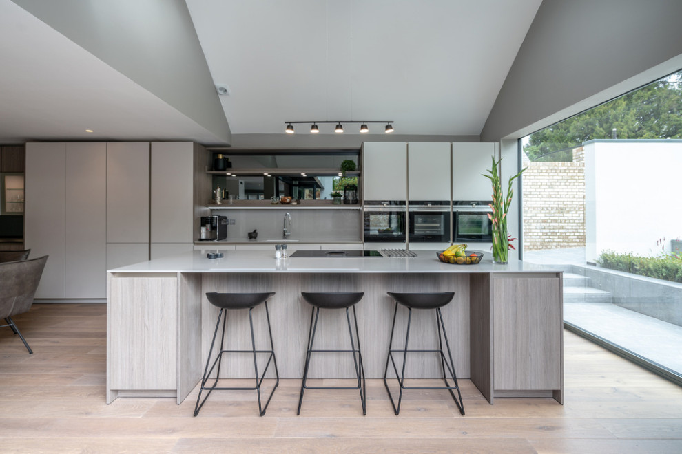 Design ideas for a large contemporary eat-in kitchen in Hampshire with a single-bowl sink, flat-panel cabinets, grey cabinets, grey splashback, black appliances, light hardwood floors, with island and grey benchtop.