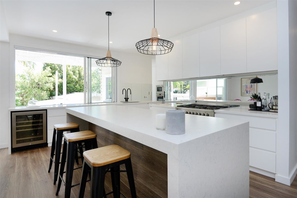 Beach style l-shaped eat-in kitchen in Sunshine Coast with an undermount sink, flat-panel cabinets, white cabinets, mirror splashback, stainless steel appliances, medium hardwood floors, with island, brown floor and white benchtop.