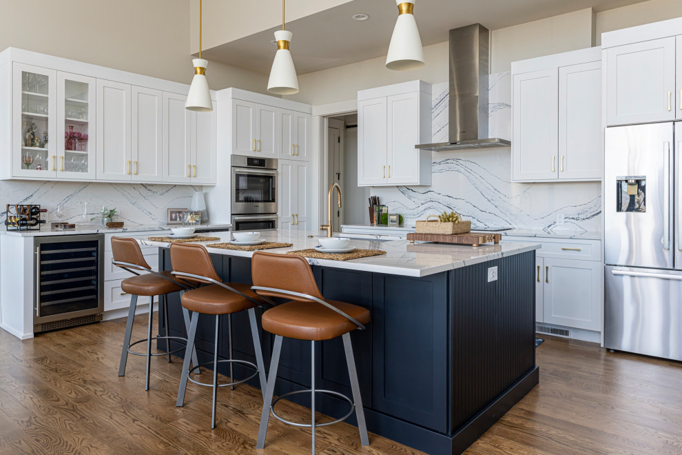 Transitional l-shaped medium tone wood floor and brown floor kitchen photo with an undermount sink, shaker cabinets, white cabinets, white backsplash, stone slab backsplash, stainless steel appliances, an island and white countertops
