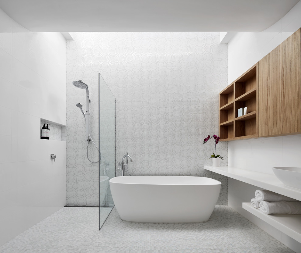 Photo of a mid-sized contemporary master bathroom in Melbourne with furniture-like cabinets, brown cabinets, a freestanding tub, an open shower, a wall-mount toilet, multi-coloured tile, mosaic tile, white walls, mosaic tile floors, a vessel sink, solid surface benchtops, white floor, an open shower and white benchtops.