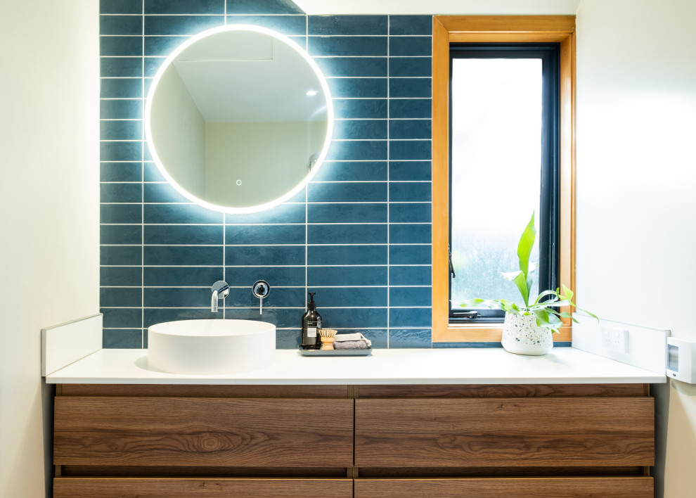 На фото: главная ванная комната среднего размера в стиле модернизм с фасадами разных видов, фасадами цвета дерева среднего тона, угловым душем, унитазом-моноблоком, синей плиткой, керамической плиткой, белыми стенами, полом из керамической плитки, настольной раковиной, столешницей из искусственного камня, серым полом, душем с раздвижными дверями, белой столешницей, тумбой под одну раковину и подвесной тумбой