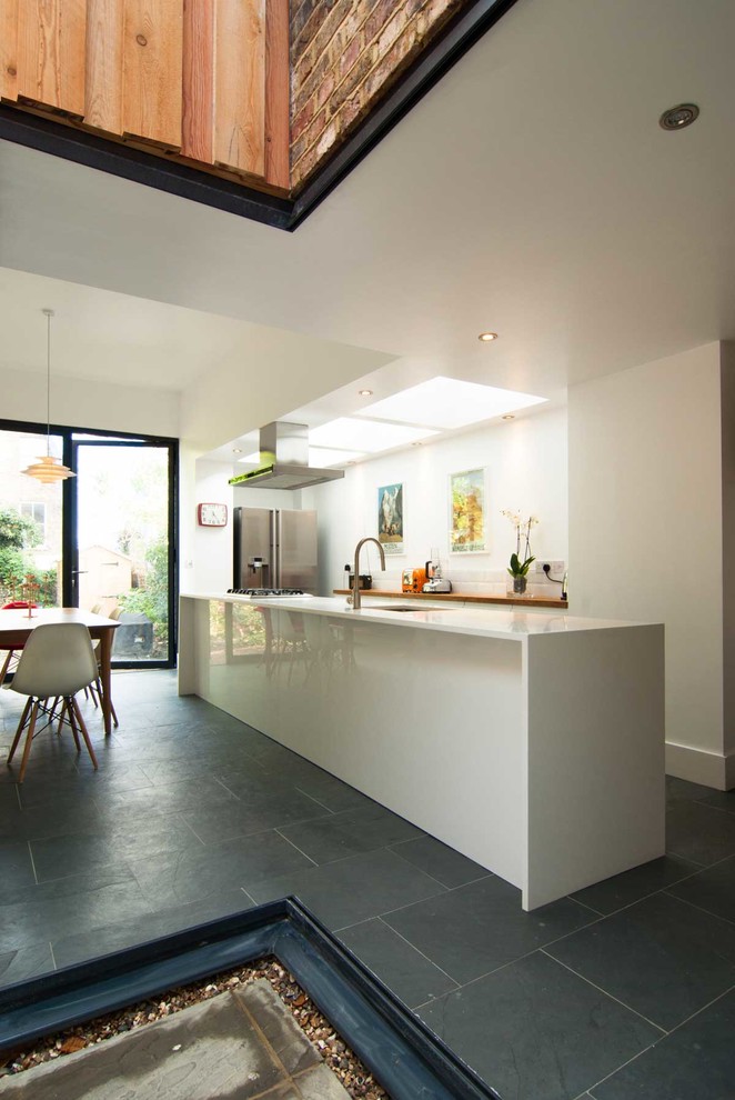 Mid-sized contemporary single-wall eat-in kitchen in London with a single-bowl sink, flat-panel cabinets, white cabinets, wood benchtops, white splashback, ceramic splashback, stainless steel appliances, slate floors and with island.