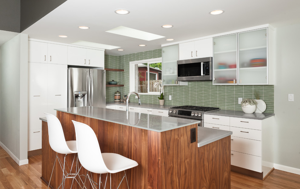 This is an example of a mid-sized midcentury l-shaped eat-in kitchen in Portland with flat-panel cabinets, medium wood cabinets, quartz benchtops, green splashback, glass tile splashback, stainless steel appliances, medium hardwood floors, with island, brown floor and grey benchtop.