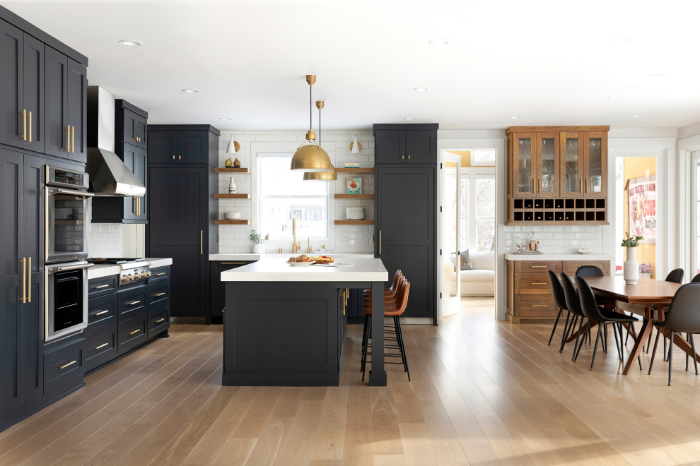 This is an example of a transitional eat-in kitchen in Minneapolis with a farmhouse sink, blue cabinets, quartz benchtops, white splashback, ceramic splashback, stainless steel appliances, light hardwood floors, with island, brown floor and white benchtop.