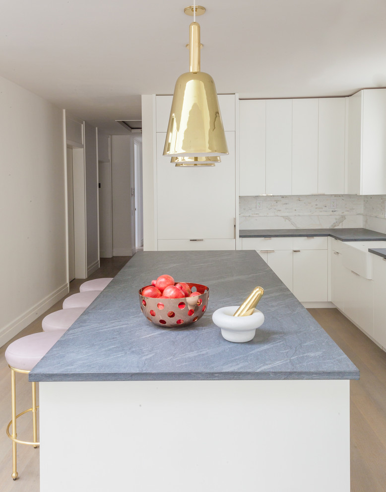 Inspiration for a mid-sized modern l-shaped eat-in kitchen in New York with a farmhouse sink, flat-panel cabinets, white cabinets, white splashback, stone tile splashback, white appliances, light hardwood floors, with island, granite benchtops and grey benchtop.