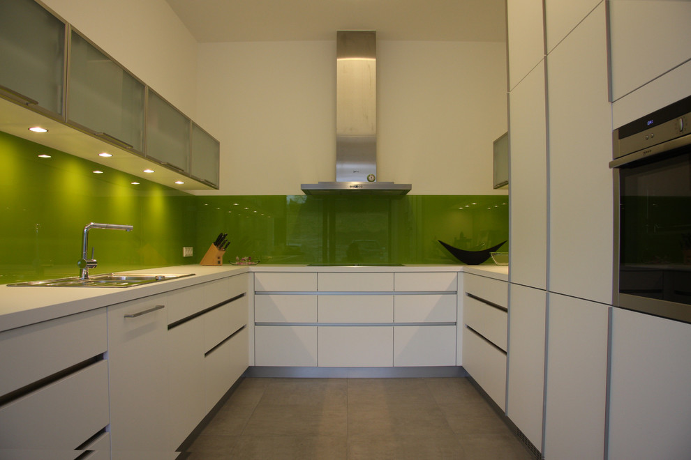 Mid-sized contemporary u-shaped separate kitchen in Dusseldorf with a drop-in sink, flat-panel cabinets, white cabinets, green splashback, glass sheet splashback, stainless steel appliances and no island.
