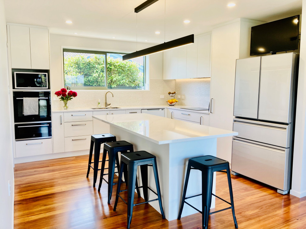 Photo of a medium sized contemporary l-shaped kitchen/diner in Christchurch with a double-bowl sink, white splashback, ceramic splashback, stainless steel appliances, an island, brown floors, flat-panel cabinets, white cabinets, engineered stone countertops, medium hardwood flooring and white worktops.