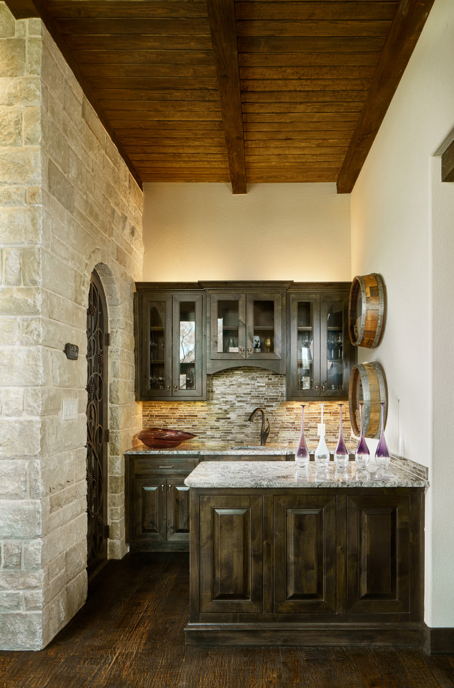 Photo of a large traditional wet bar in Other with an undermount sink, raised-panel cabinets, dark wood cabinets, granite benchtops, multi-coloured splashback, glass tile splashback, dark hardwood floors and brown floor.