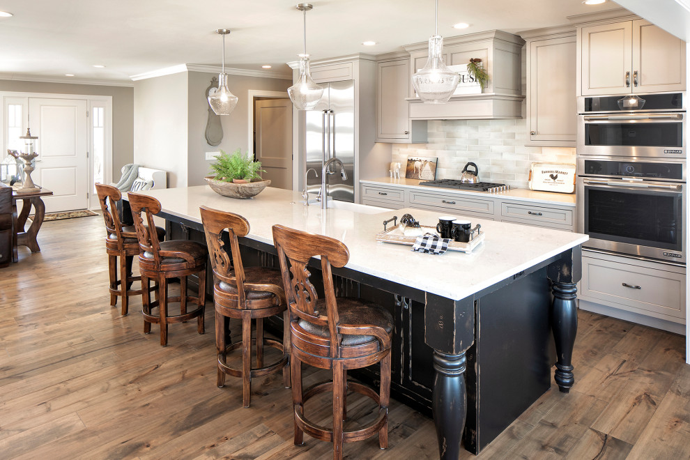 This is an example of a large country open plan kitchen in Minneapolis with a farmhouse sink, ceramic splashback, stainless steel appliances, medium hardwood floors, with island, brown floor, white benchtop, granite benchtops, shaker cabinets, multi-coloured splashback and grey cabinets.