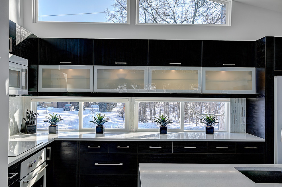 Inspiration for a mid-sized contemporary l-shaped open plan kitchen in Grand Rapids with an undermount sink, glass-front cabinets, quartz benchtops, white splashback, glass tile splashback, white appliances, light hardwood floors, with island and black cabinets.