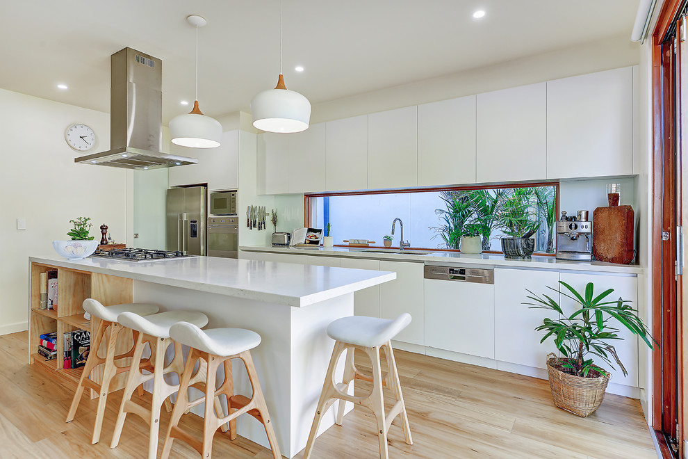 Design ideas for a large beach style galley open plan kitchen in Sydney with flat-panel cabinets, white cabinets, stainless steel appliances, light hardwood floors, brown floor, white benchtop, an undermount sink, quartz benchtops, white splashback, glass sheet splashback and with island.