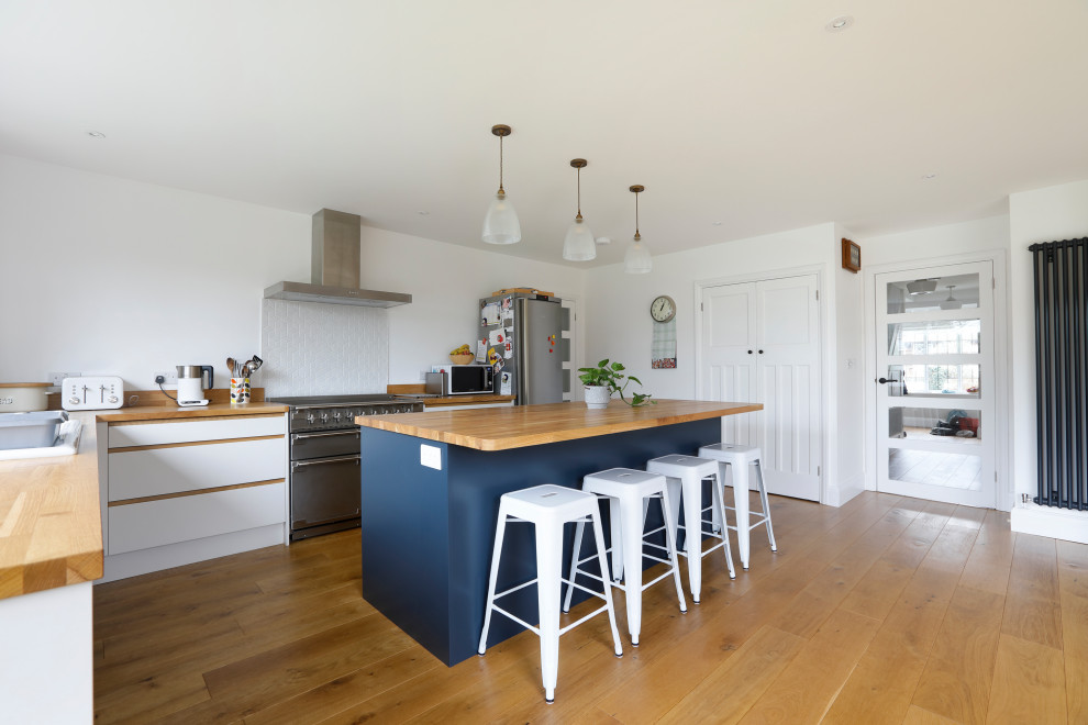 This is an example of a large contemporary l-shaped kitchen/diner in Sussex with a double-bowl sink, flat-panel cabinets, white cabinets, wood worktops, white splashback, ceramic splashback, stainless steel appliances, medium hardwood flooring, an island, brown floors and brown worktops.