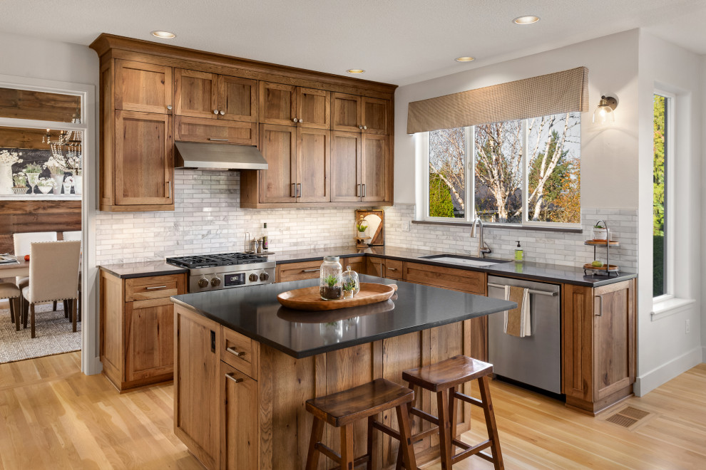 Design ideas for a mid-sized country l-shaped eat-in kitchen in Portland with an undermount sink, shaker cabinets, medium wood cabinets, granite benchtops, stainless steel appliances, light hardwood floors, with island, grey benchtop, white splashback, marble splashback and brown floor.