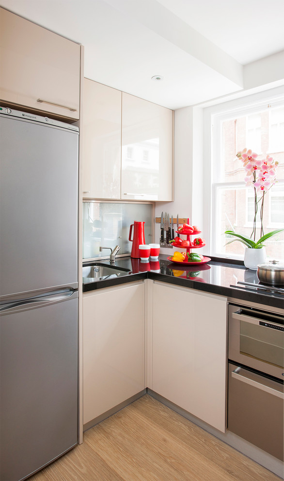 This is an example of a small contemporary u-shaped enclosed kitchen in London with a built-in sink, flat-panel cabinets, beige cabinets, granite worktops, glass sheet splashback, stainless steel appliances, medium hardwood flooring, no island and brown floors.