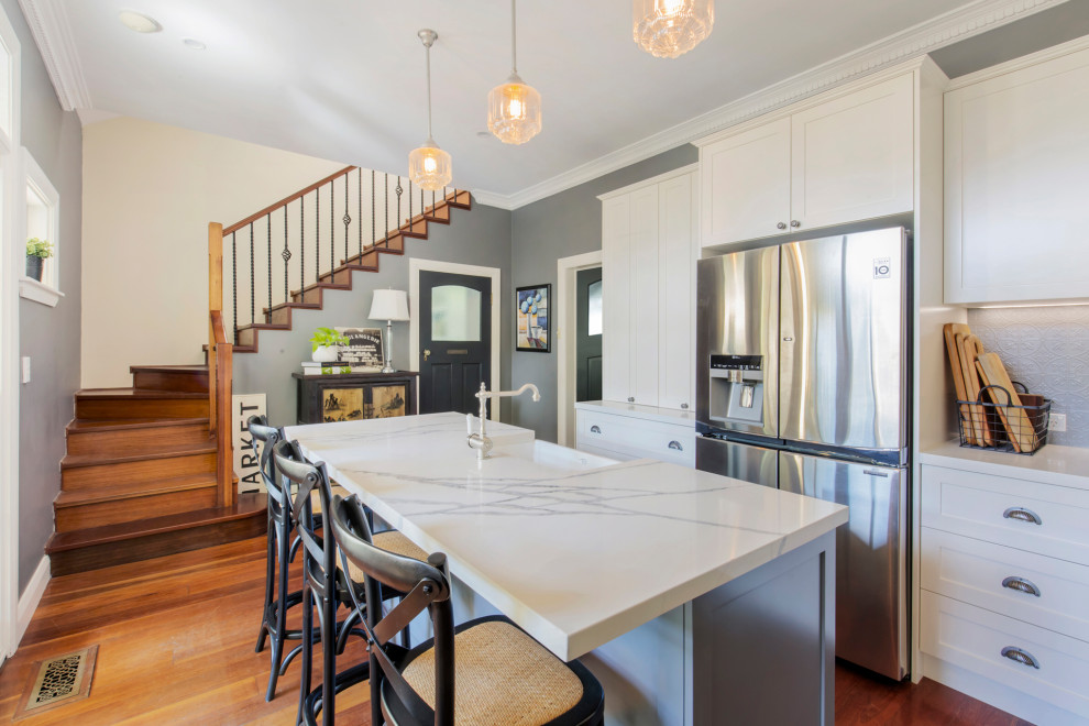 Inspiration for a mid-sized traditional l-shaped eat-in kitchen in Sydney with a farmhouse sink, shaker cabinets, white cabinets, quartz benchtops, grey splashback, metal splashback, black appliances, medium hardwood floors, with island, brown floor and multi-coloured benchtop.