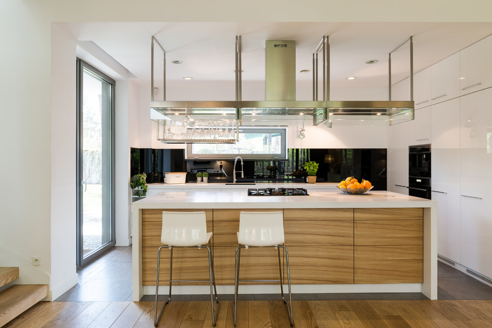 This is an example of a mid-sized modern galley eat-in kitchen in Los Angeles with a drop-in sink, flat-panel cabinets, medium wood cabinets, solid surface benchtops, black splashback, mirror splashback, black appliances, medium hardwood floors, with island, multi-coloured floor and white benchtop.