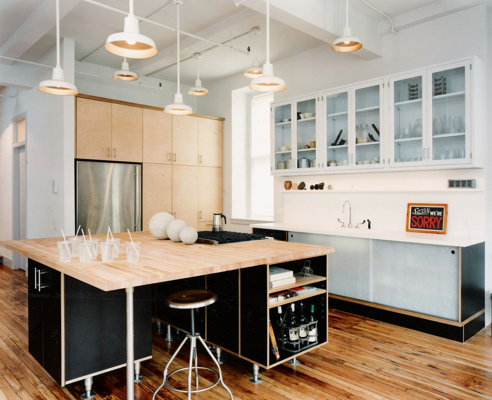 This is an example of an industrial kitchen in New York with glass-front cabinets, solid surface benchtops, white splashback, stainless steel appliances, with island, light wood cabinets and medium hardwood floors.