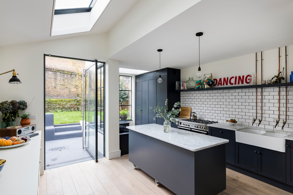 Contemporary galley open plan kitchen in London with a farmhouse sink, shaker cabinets, black cabinets, white splashback, subway tile splashback, stainless steel appliances, light hardwood floors, with island and beige floor.
