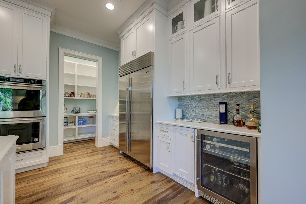 Foto på ett mellanstort maritimt vit kök, med en undermonterad diskho, skåp i shakerstil, vita skåp, marmorbänkskiva, blått stänkskydd, stänkskydd i glaskakel, rostfria vitvaror, mellanmörkt trägolv, en köksö och brunt golv