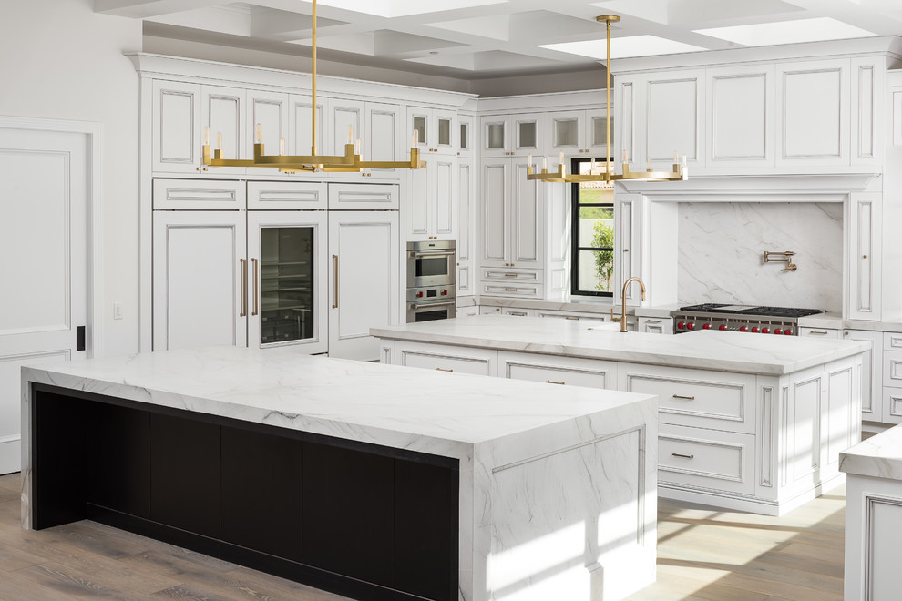 This is an example of a transitional u-shaped kitchen in Phoenix with a farmhouse sink, recessed-panel cabinets, white cabinets, white splashback, stone slab splashback, panelled appliances, light hardwood floors, multiple islands, beige floor and white benchtop.