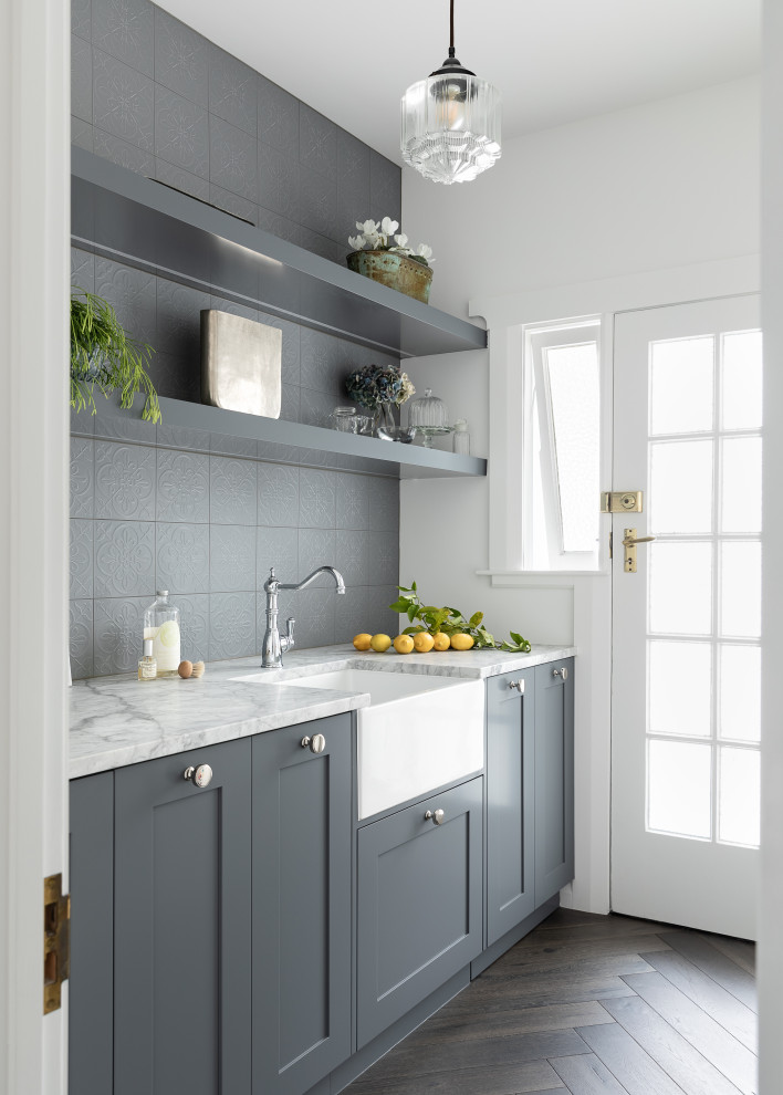 This is an example of a small transitional galley dedicated laundry room in Auckland with a farmhouse sink, shaker cabinets, grey cabinets, quartzite benchtops, grey splashback, ceramic splashback, grey walls, dark hardwood floors, a concealed washer and dryer, brown floor, multi-coloured benchtop, recessed and decorative wall panelling.