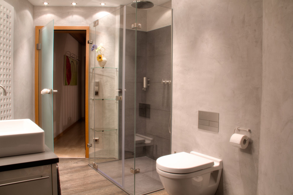 Photo of a mid-sized contemporary 3/4 bathroom in Munich with shaker cabinets, grey cabinets, a curbless shower, a wall-mount toilet, gray tile, grey walls, laminate floors, a vessel sink, wood benchtops, brown floor, a hinged shower door, grey benchtops, a single vanity and a freestanding vanity.