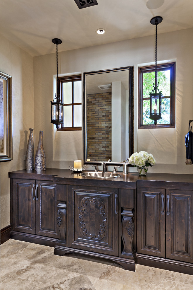 Inspiration for a large mediterranean powder room in Phoenix with raised-panel cabinets, dark wood cabinets, white tile, white walls, travertine floors, a drop-in sink, wood benchtops, beige floor, brown benchtops and a one-piece toilet.