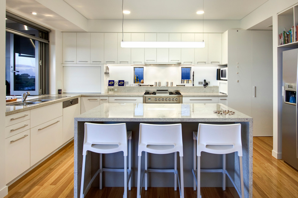 Large contemporary l-shaped open plan kitchen in Brisbane with a drop-in sink, white splashback, glass sheet splashback, stainless steel appliances, with island, flat-panel cabinets, white cabinets, light hardwood floors and brown floor.