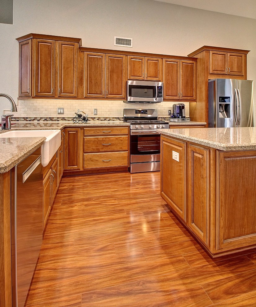 Example of a mid-sized l-shaped medium tone wood floor eat-in kitchen design in Phoenix with a farmhouse sink, raised-panel cabinets, medium tone wood cabinets, beige backsplash, stone tile backsplash, stainless steel appliances and an island