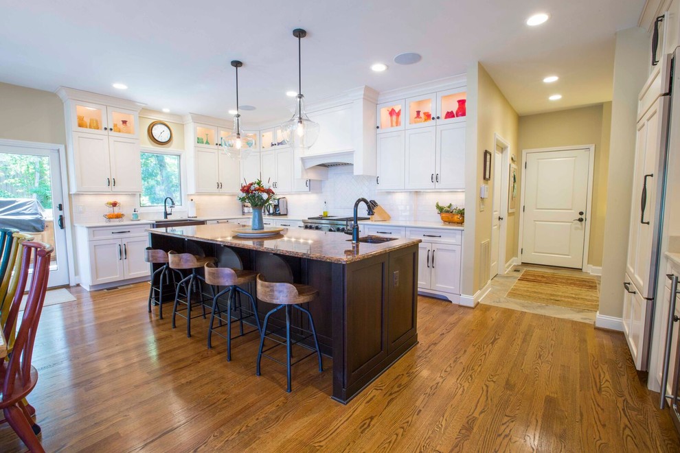 Inspiration for a large transitional l-shaped light wood floor and brown floor enclosed kitchen remodel with a farmhouse sink, shaker cabinets, white cabinets, solid surface countertops, white backsplash, porcelain backsplash, stainless steel appliances and an island