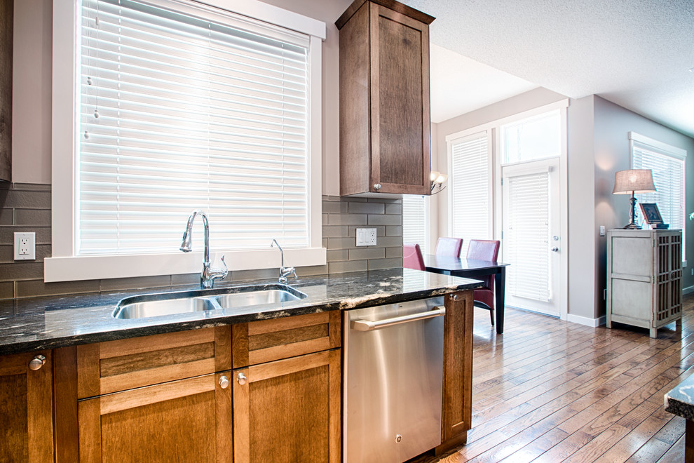 Example of a mid-sized mountain style u-shaped medium tone wood floor and brown floor eat-in kitchen design in Calgary with an undermount sink, shaker cabinets, medium tone wood cabinets, granite countertops, gray backsplash, glass tile backsplash, stainless steel appliances and an island
