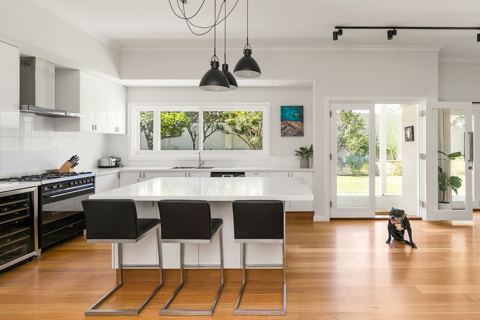 Design ideas for a contemporary l-shaped eat-in kitchen in Perth with a double-bowl sink, flat-panel cabinets, white cabinets, white splashback, black appliances, medium hardwood floors, with island, brown floor and white benchtop.