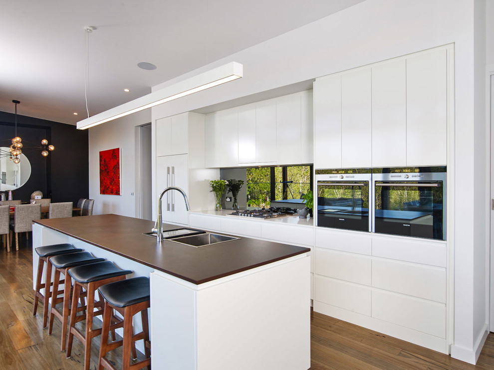 Large contemporary galley kitchen pantry in Other with a drop-in sink, flat-panel cabinets, white cabinets, solid surface benchtops, metallic splashback, mirror splashback, stainless steel appliances, light hardwood floors, with island, brown floor and brown benchtop.