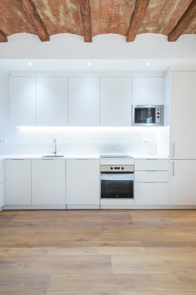 This is an example of a medium sized contemporary l-shaped open plan kitchen in Barcelona with a single-bowl sink, flat-panel cabinets, white cabinets, white splashback, stainless steel appliances, medium hardwood flooring, no island, brown floors and white worktops.