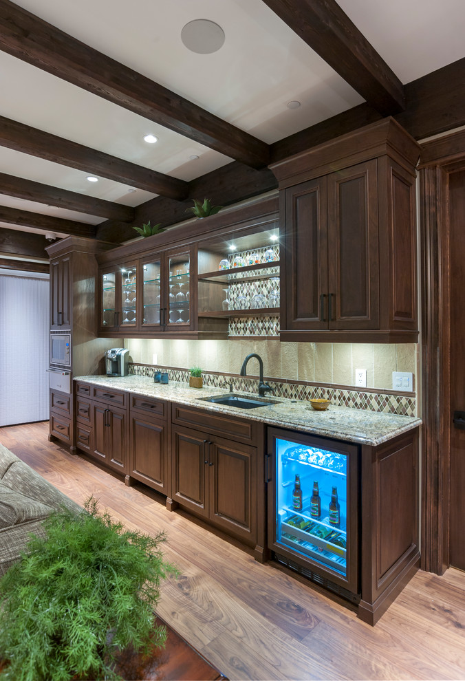 This is an example of a mid-sized country single-wall wet bar in Calgary with an undermount sink, raised-panel cabinets, granite benchtops, beige splashback, stone tile splashback, medium hardwood floors, brown floor and dark wood cabinets.