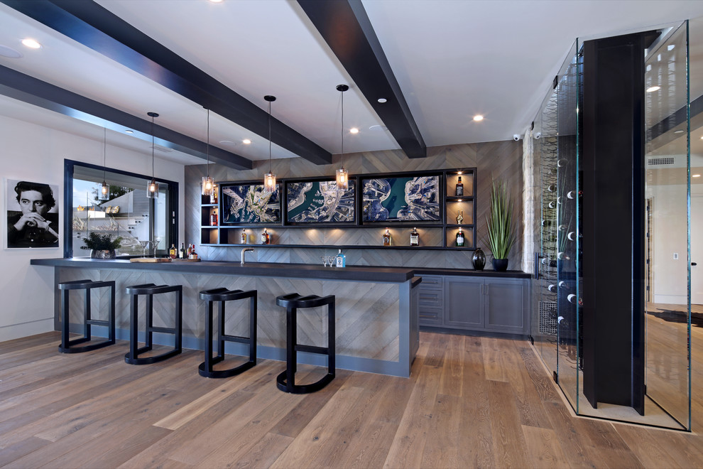 Photo of a contemporary galley wet bar in Los Angeles with shaker cabinets, grey cabinets, grey splashback, timber splashback, medium hardwood floors, brown floor and black benchtop.