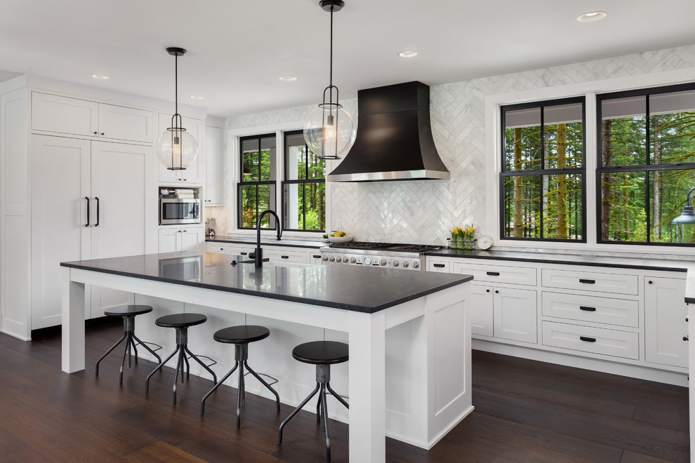 This is an example of a mid-sized transitional u-shaped eat-in kitchen in DC Metro with a farmhouse sink, white cabinets, quartz benchtops, white splashback, marble splashback, stainless steel appliances, dark hardwood floors, with island, brown floor, black benchtop and shaker cabinets.