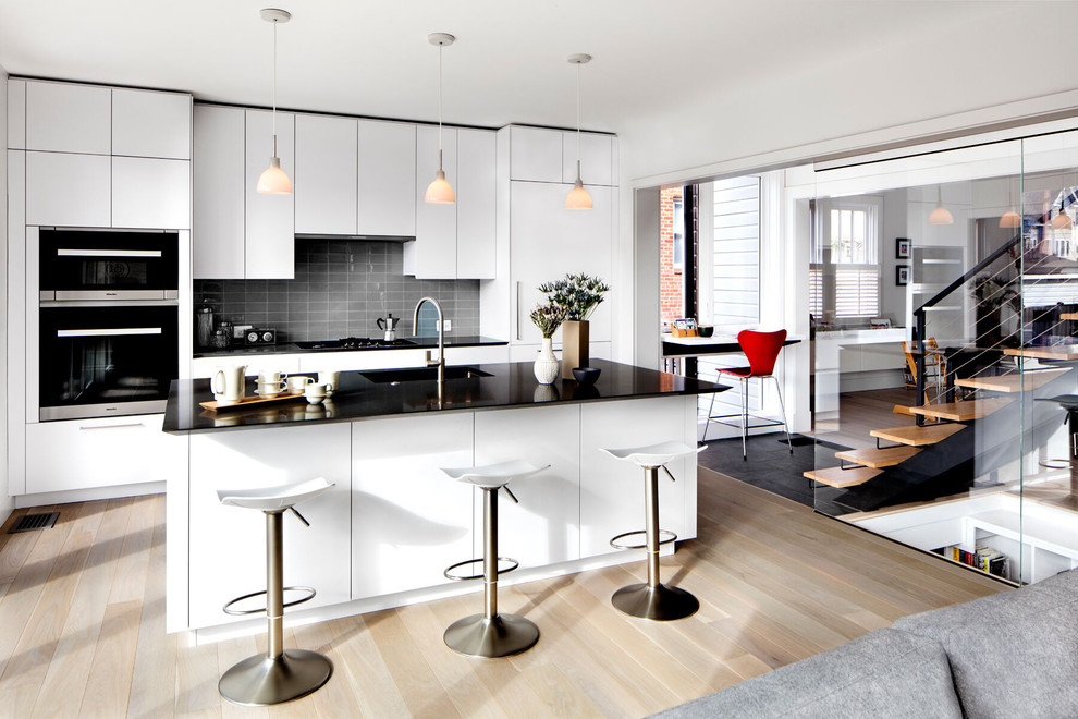 This is an example of a modern eat-in kitchen in DC Metro with an undermount sink, flat-panel cabinets, white cabinets, quartz benchtops, grey splashback, subway tile splashback, stainless steel appliances, light hardwood floors and with island.