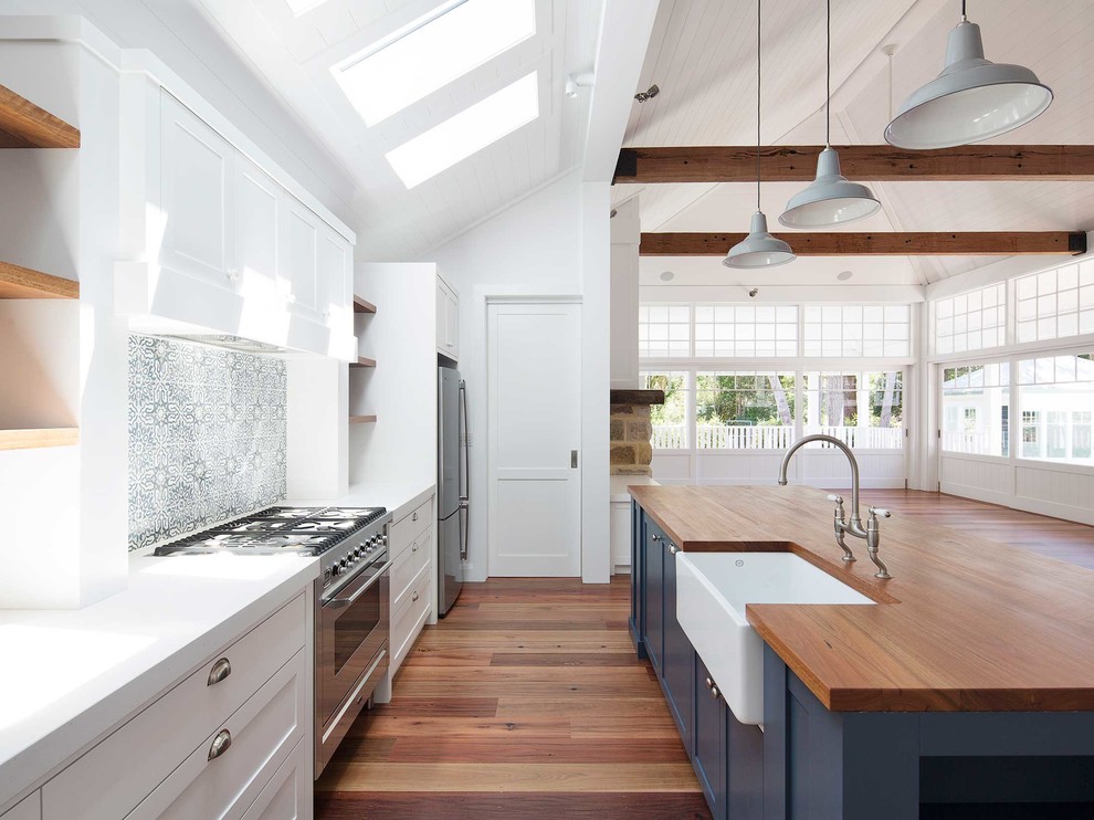 This is an example of a large beach style galley open plan kitchen in Sydney with a farmhouse sink, shaker cabinets, blue cabinets, wood benchtops, multi-coloured splashback, cement tile splashback, stainless steel appliances, medium hardwood floors, with island and brown floor.