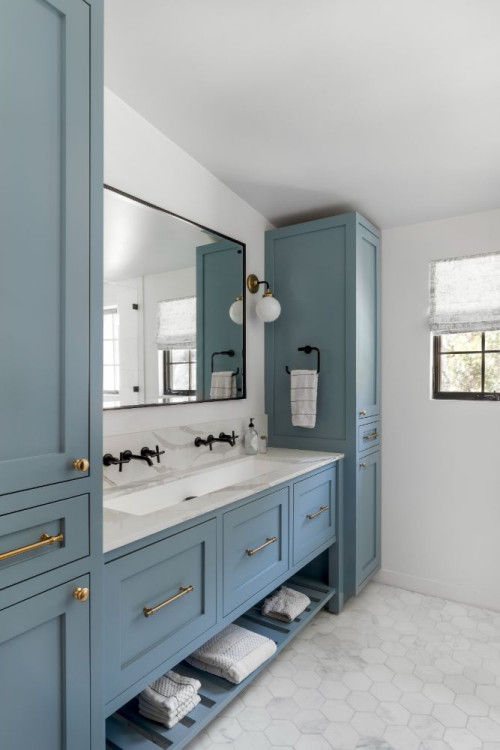 Elegant bathroom with blue shaker-style cabinets, brass hardware, double-sink vanity, large mirror, and open shelving for towels on a white hexagon tile floor.