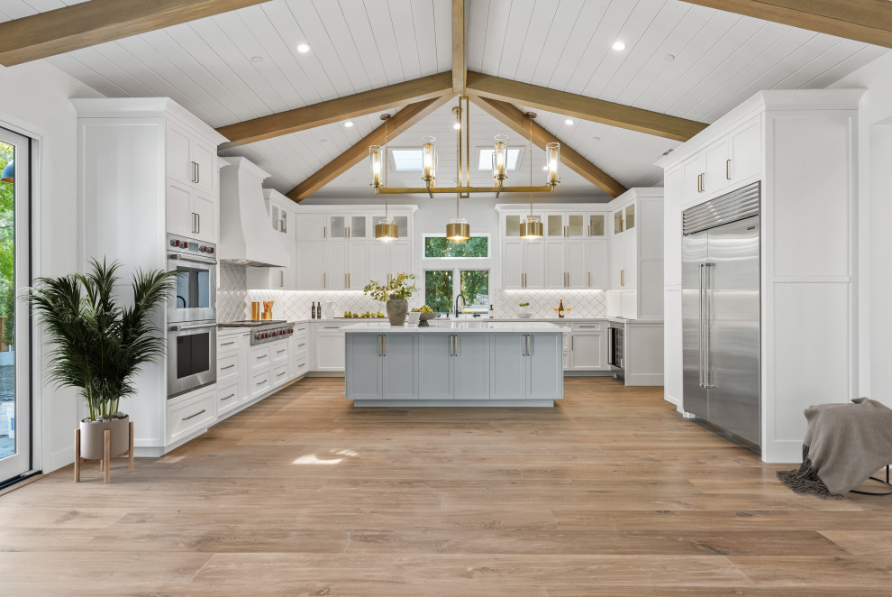 Inredning av ett modernt mycket stort vit vitt kök, med en rustik diskho, luckor med infälld panel, vita skåp, bänkskiva i kvartsit, vitt stänkskydd, stänkskydd i porslinskakel, rostfria vitvaror, ljust trägolv, en köksö och brunt golv