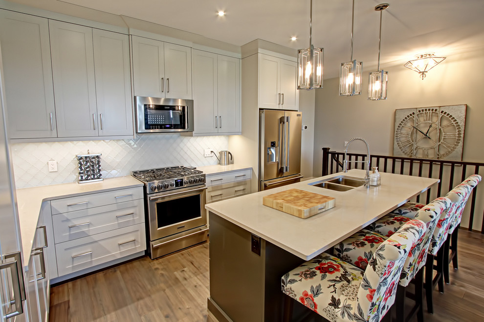 Mid-sized transitional l-shaped medium tone wood floor and brown floor open concept kitchen photo in Toronto with a double-bowl sink, shaker cabinets, white cabinets, quartz countertops, white backsplash, glass tile backsplash, stainless steel appliances, an island and beige countertops