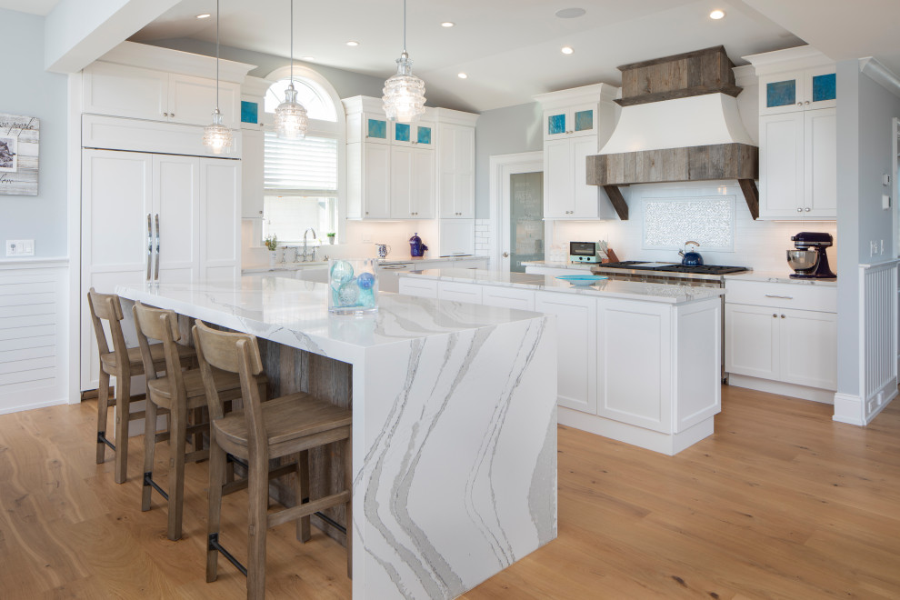 Inspiration for a large beach style l-shaped eat-in kitchen in New York with a farmhouse sink, shaker cabinets, white cabinets, quartz benchtops, white splashback, subway tile splashback, stainless steel appliances, light hardwood floors, multiple islands, beige floor and white benchtop.