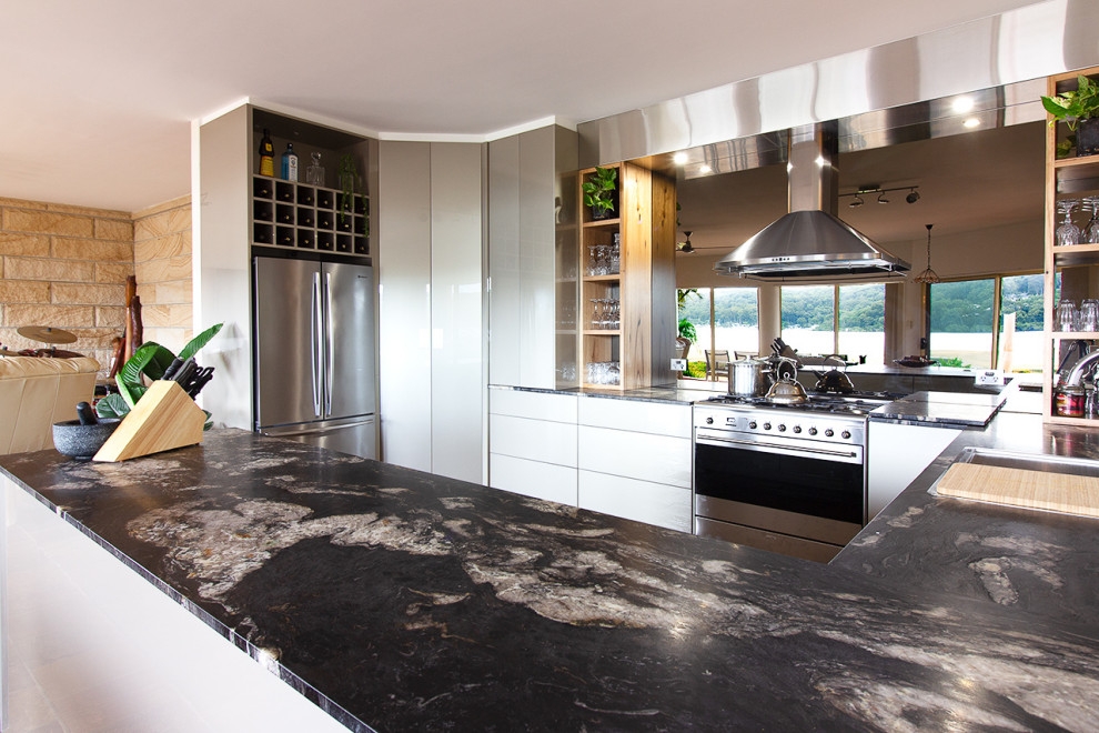 Inspiration for a large modern u-shaped kitchen pantry in Central Coast with a double-bowl sink, flat-panel cabinets, beige cabinets, granite benchtops, metallic splashback, glass sheet splashback, stainless steel appliances, slate floors, a peninsula, brown floor and black benchtop.