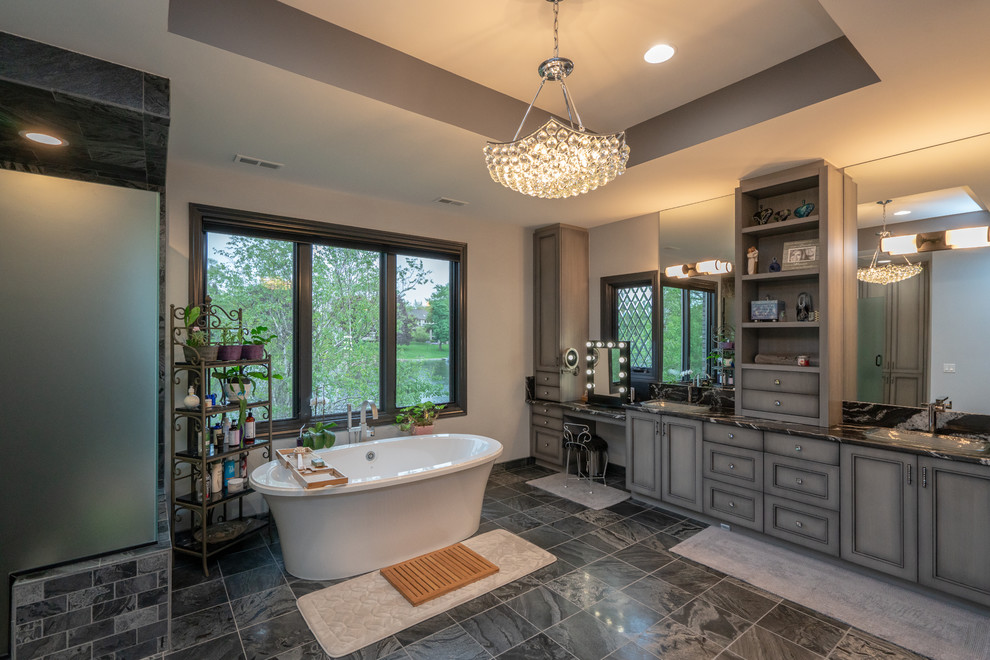 This is an example of a large traditional master bathroom in Detroit with beaded inset cabinets, grey cabinets, a freestanding tub, a corner shower, gray tile, stone tile, grey walls, porcelain floors, an undermount sink, granite benchtops, black floor, a hinged shower door and black benchtops.