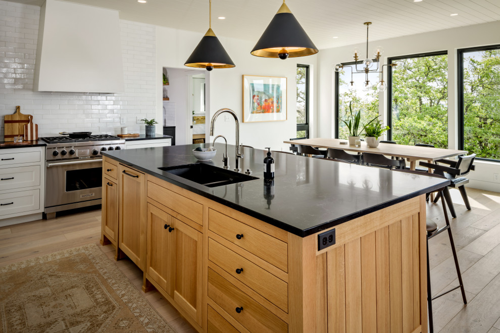 This is an example of a mid-sized transitional eat-in kitchen in Other with shaker cabinets, medium wood cabinets, white splashback, stainless steel appliances, light hardwood floors and black benchtop.