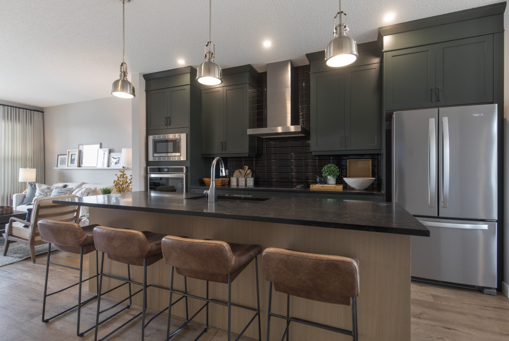This is an example of a mid-sized country kitchen in Calgary with shaker cabinets, black splashback, stainless steel appliances, with island, brown floor and black benchtop.