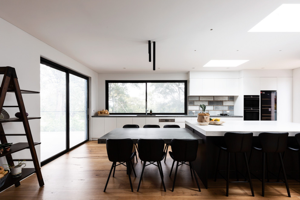 Design ideas for a contemporary galley kitchen in Sydney with flat-panel cabinets, white cabinets, grey splashback, black appliances, medium hardwood floors, with island, brown floor and white benchtop.